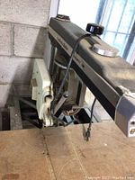 Close-up of the radial arm saw showing the blade and arm mechanism on a wooden surface in a garage.
