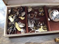 Top view inside case showing assorted cufflinks, tie clips, and pins in metal tones of gold and silver, some with decorative elements like birds or geometric shapes.