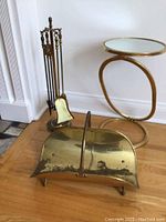 Photo showing brass fireplace tool set on stand, brass firewood carrier, and small round accent table with mirrored top on hardwood floor.