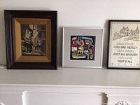 Front view of the three framed art pieces placed side by side on a mantel, showing the oil painting, ceramic tile, and embroidered linen.