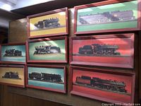 Eight framed prints of various railroad steam engines, arranged in two columns on the floor, each framed in reddish-brown wood, colorful backgrounds in yellow, green, and red.