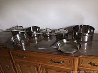 Full overview of the Logostina stainless steel cookware set showing pots, pans, and lids arranged on a wooden surface.
