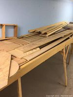View of several plywood sheets, curved panels, and boards stacked on a wooden folding sawhorse table against a light blue wall.
