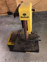 Front view of the yellow Rockwell bench grinder showing the sanding belt arm and work platform.