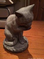 Stone statue of a cat sitting on a round base, angled front view showing head bowed and detailed texture.