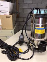 Photo showing full view of stainless steel sump pump including long power cable and plug on a table surface.