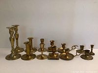 Wide view of all 13 brass candlesticks showing variety of sizes and styles from tall to short chamber pieces.
