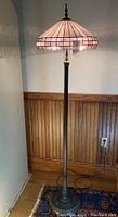 Full view of the tall brass floor lamp with its stained glass dome shade near a wooden paneled wall, showing overall shape and height.