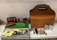 Photo showing a brown leather carrying case, sunglasses, eyeglass case, reading materials, pocket knives, lighters, and keys spread out on a shelf.