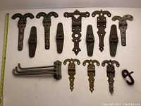Wide view of all twelve metal hinges and hardware including several decorative strap hinges, butt hinges, folding extension arms, and a metal hook on white surface with measuring tape.