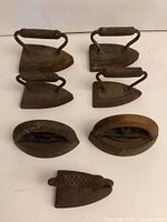 Top view of seven vintage cast iron irons showing various sizes and shapes with textured handles.