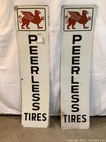Front view of two vertical Peerless Tires sheet metal signs with red griffin emblems and vertical black text on white background, showing paint wear.
