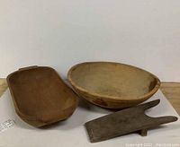 Photo showing the set of three primitive wooden items: oblong bowl, round bowl, and boot pull tool, all placed on white surface against white backdrop.
