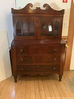 Front view of late 19th century wooden secretary desk showing upper cabinet with glass doors and ornate woodwork, lower section with three drawers and turned feet.