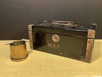Front view of black metal strong box with reinforced corners, hallmark 'MRP' below lock, and metal handle on top.