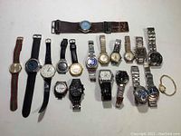 Overview photo showing 20 wristwatches arranged in a row on a white background. Mixed styles of men's and women's watches with various band materials and dial colors.