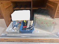 Photo shows open wooden cabinet with plastic baskets containing batteries, light bulbs, and two vintage wooden metre sticks placed in front. Also visible are plastic file folders and three magazine holders behind the baskets and sticks. Adjustable closet rod rods placed at front.