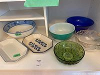 Photo showing assortment of ceramic and glass bowls and trays on white shelf, including large ceramic bowl, two ceramic trays, and various colored glass bowls.