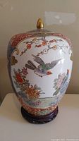 Front view of the urn showing detailed bird and floral designs on white ceramic with red and gold borders, including the wooden stand beneath.