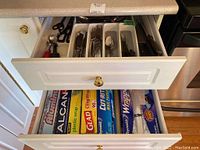 View of two drawers: top drawer with cutlery organized in white tray; bottom drawer with rolls of plastic wrap, wax paper, aluminum foil, and foil wrap