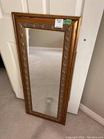 Full view of framed mirror resting against door, showing bevelled edge and metallic distressed ornate inner border on wooden frame