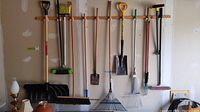 Wide view of all garden tools hung on an attached wooden rack on a garage wall, including shovels, rakes, brooms, and garden edger, showing item variety and condition.