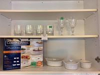 Photo showing boxed Corningware French White 8-piece set with three white ceramic dishes out of the box, and 4 glass tumblers with 2 stemmed wine glasses on shelf above.