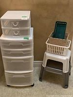 View of two plastic drawer units stacked, and step stools with organizing bins on top against a wall
