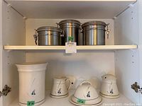 Photo showing three stainless steel canisters on top shelf and pottery items on lower shelf including utensil holder, creamer, sugar bowl, and butter dish.