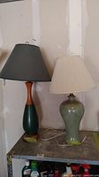 Photo showing both lamps side by side on a table with light background; left lamp has wood and green painted base with dark green shade, right lamp has pale green ceramic base with cream pleated shade.