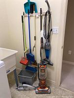 Photo showing upright Shark vacuum and handheld Shark vacuum on the floor with brooms, dustpans and bucket hanging or placed alongside wall