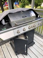 Front and left side of the stainless steel Nexgrill propane grill with cover partially over the top, showing multiple control knobs and cabinet doors open below the grill area.