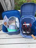 Two blue plastic totes filled with barbecue and picnic accessories viewed from above showing mixed contents including grill basket, BBQ tools, and plastic kitchenware.