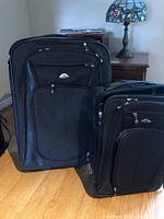 Photo showing two black Samsonite suitcases, one larger and one smaller, standing upright on wood floor near furniture.