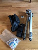 Photo showing aluminum tripod, black leather-like camera case, black nylon fanny pack, and plastic bag with assorted used stamps on wood floor