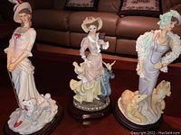 Shows all three porcelain female figurines together, each around 11-12 inches tall, detailed with dogs and birds.