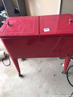 Full view of the red cooler showing hinged double lids and caster wheels under legs.