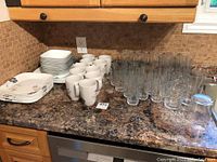 Corelle dish set arrangement showing stacked dinner plates, luncheon plates, bowls, and mugs on a counter next to glasses.