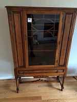 Front view of walnut cabinet showing glass door with decorative fretwork, side wood panels, key lock, lower drawer and turned legs