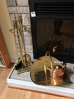 Photo showing brass fireplace tools on stand placed next to fireplace and brass wood holder with copper kettle on it.