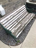 View of the full bench showing white wooden slats that are weathered and peeling, cast iron green frame, placed on gravel next to a brick wall and air conditioning unit.