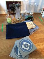 Wide view of lot items on floor including linens, crystal jug, glass vases, silver plate serving pieces, candle, Wedgwood plates boxed