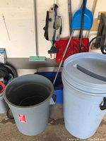 Photo of two large trash cans, one gray with no lid, the other light gray with lid and handle; shovel and cleaning tools visible in background.