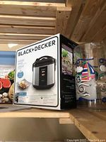 Photo of Black+Decker rice cooker in original box on wooden shelf next to decorated plastic cups
