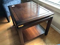 Oblique view of one wooden side table showing top surface, leg, open lower shelf, and brass accents on corners.