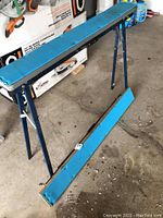 Photo showing a full view of one metal frame saw horse with blue textured DIY top attached and the other with the blue top detached and on the ground.
