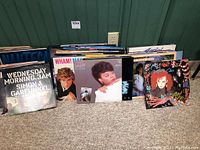 Several vinyl record albums displayed on carpet and leaning against a green paneled wall with electrical outlet visible.