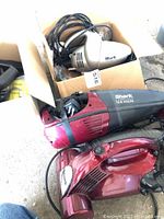 Photo showing three Shark hand vacuums including a red cordless model, a silver Original Turbo Hand Vac, and a newer red electric model in a box with cords.