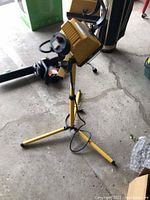 Photo showing one yellow work light mounted on a yellow folding tripod stand placed on concrete floor.