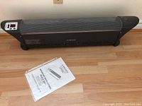 Front view of Airworks digital baseboard heater placed on hardwood floor with manual in front.